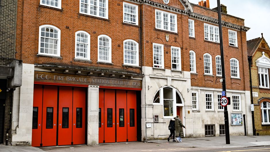 The image shows the front exterior of the LCC Fire Brigade station in Lewisham, characterized by a historic brick building with multiple white-framed sash windows. The station features prominent brigh